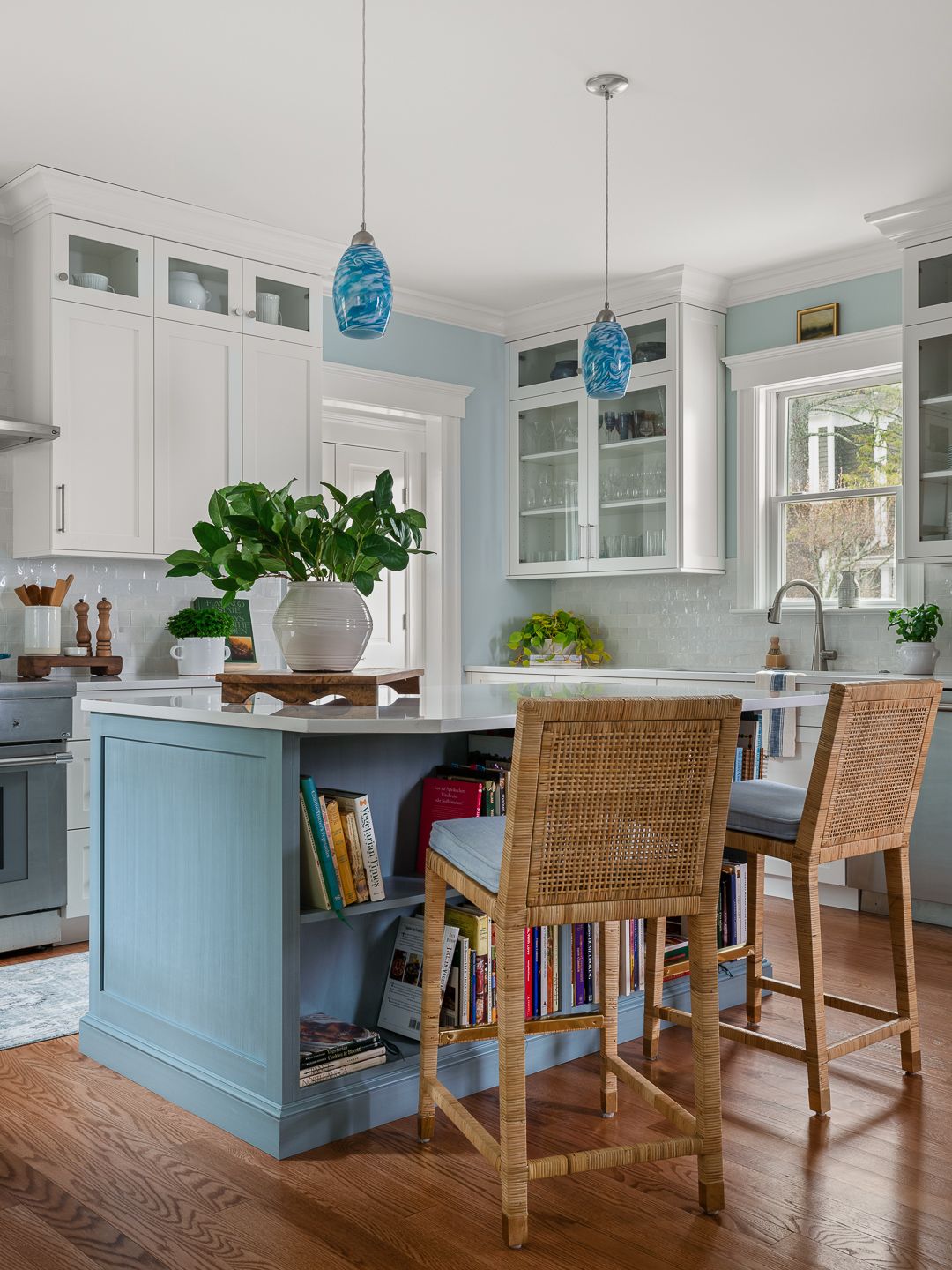 Bright and airy kitchen with white cabinetry, blue island featuring built-in cookbook shelves, rattan bar stools, and pendant lights, designed by Ginny Padula of Town & Country Design Studio and photographed by Tori Sikkema Photography.