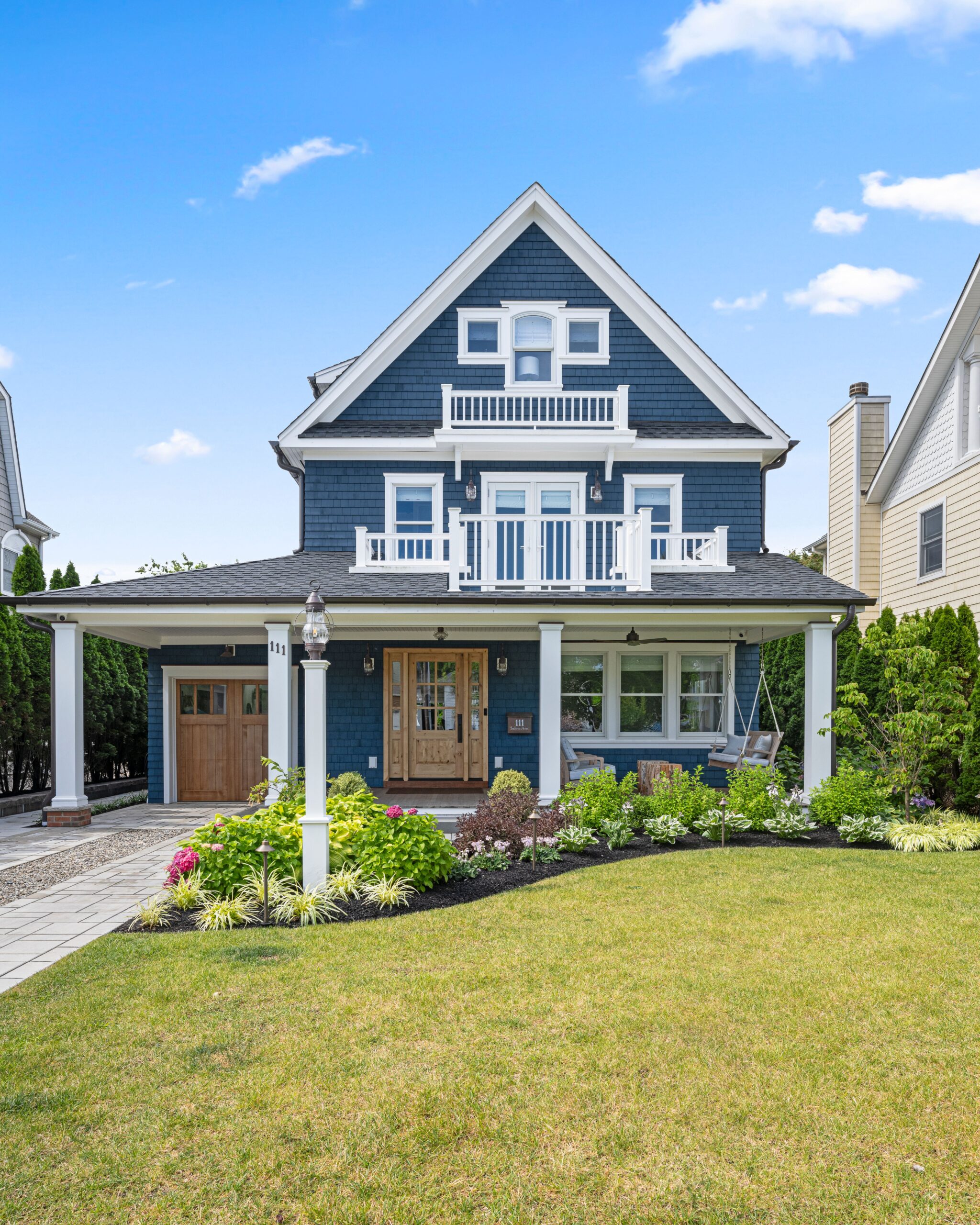 Coastal front yard with low-maintenance landscaping, featuring structured boxwoods, blooming hydrangeas, and hostas lining a paved walkway to a white beachside home with a covered porch