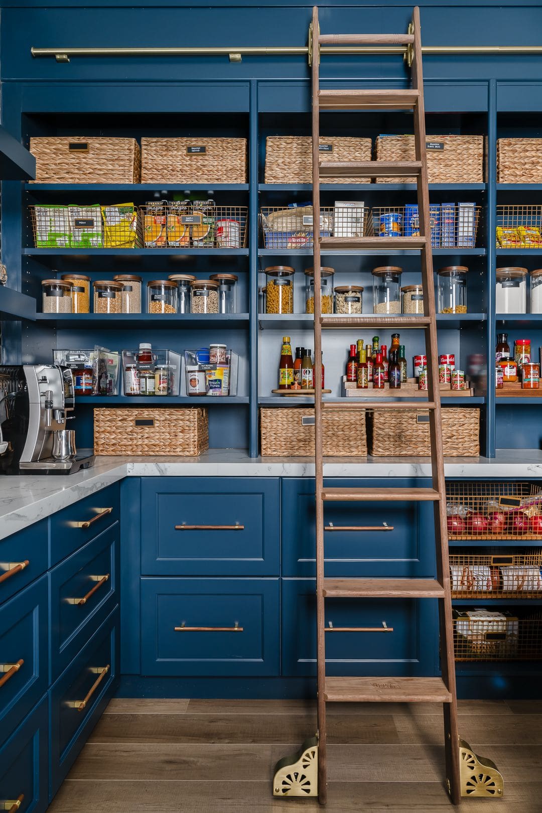 organized walk-in pantry featuring floor-to-ceiling navy cabinetry with brass hardware and a built-in brass rail supporting a rolling wooden ladder.