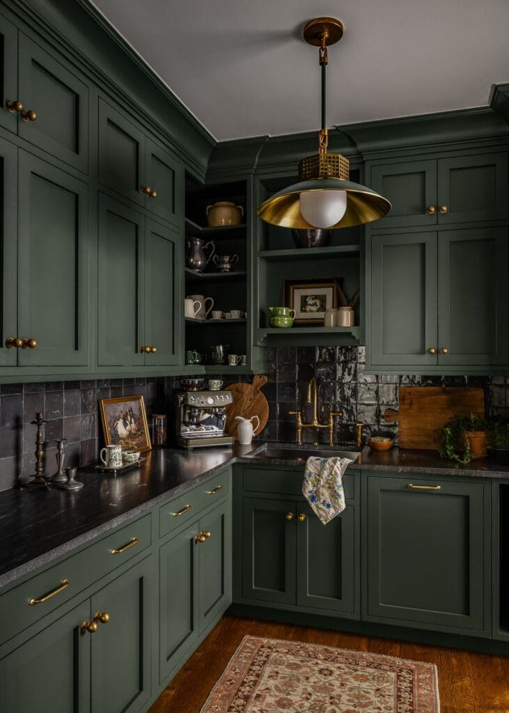 A moody, editorial-style butler’s pantry featuring deep green shaker cabinetry, aged brass hardware, and a black tiled backsplash