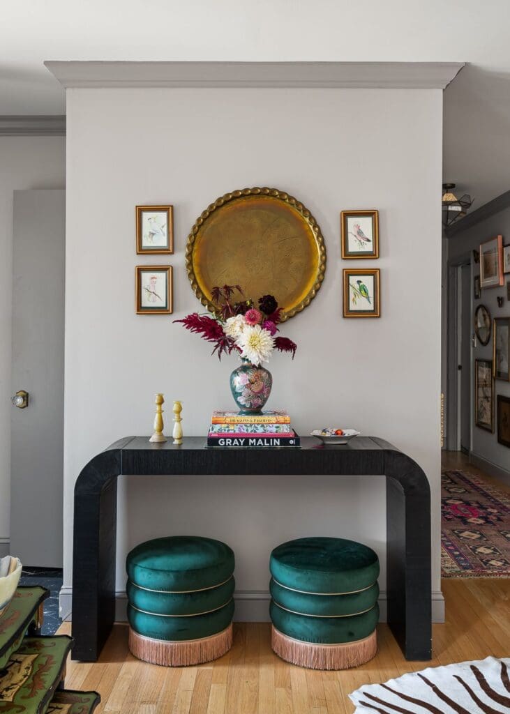A styled vignette featuring a black curved console table with a floral vase of burgundy, blush, and ivory blooms atop stacked coffee table books. Twin emerald green velvet stools with pink fringe are tucked below.