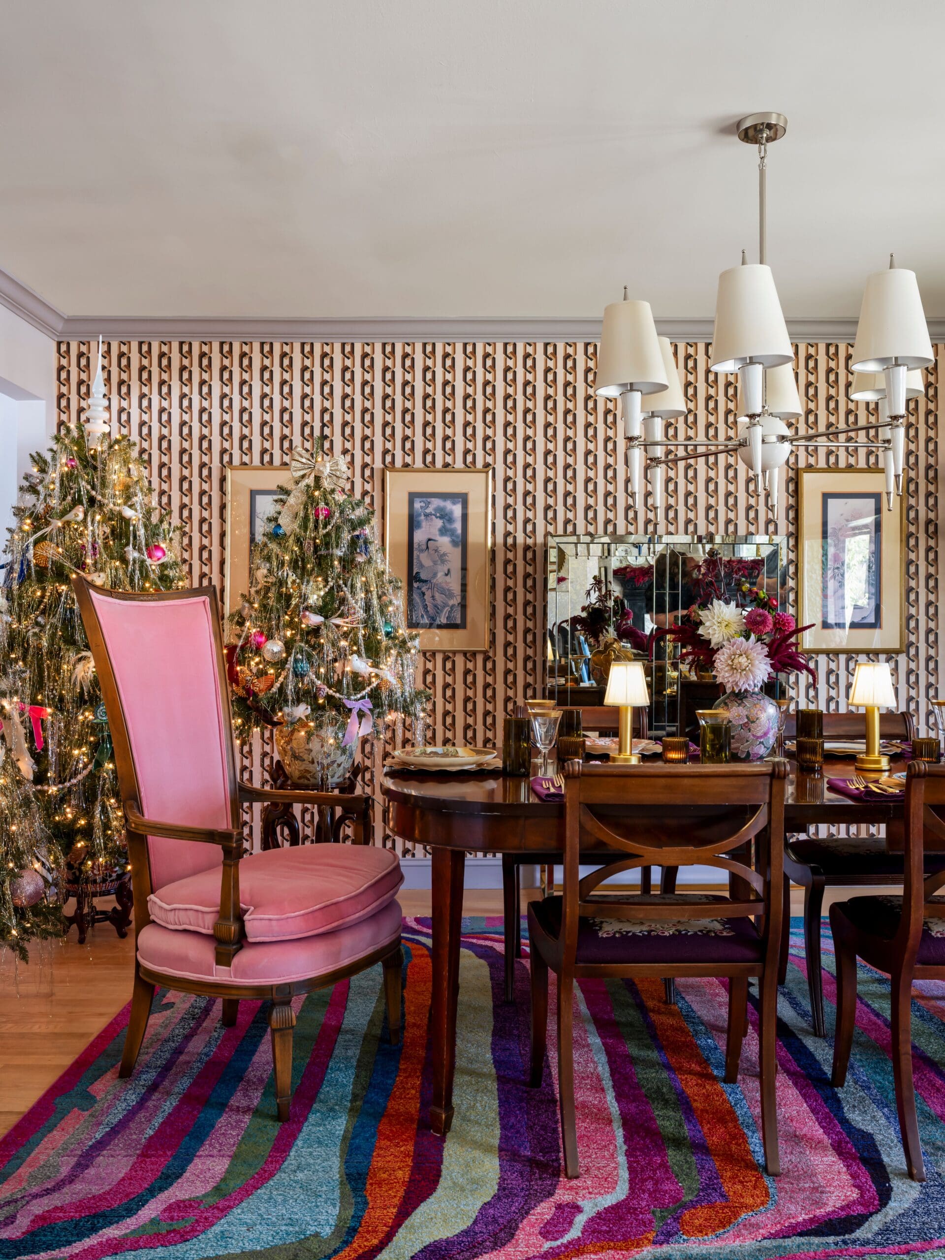 Holiday-themed dining room decorated in a maximalist style featuring two fully adorned Christmas trees with colorful ornaments, a vintage wood dining table set with glassware and floral arrangements, and a large pink high-back chair.