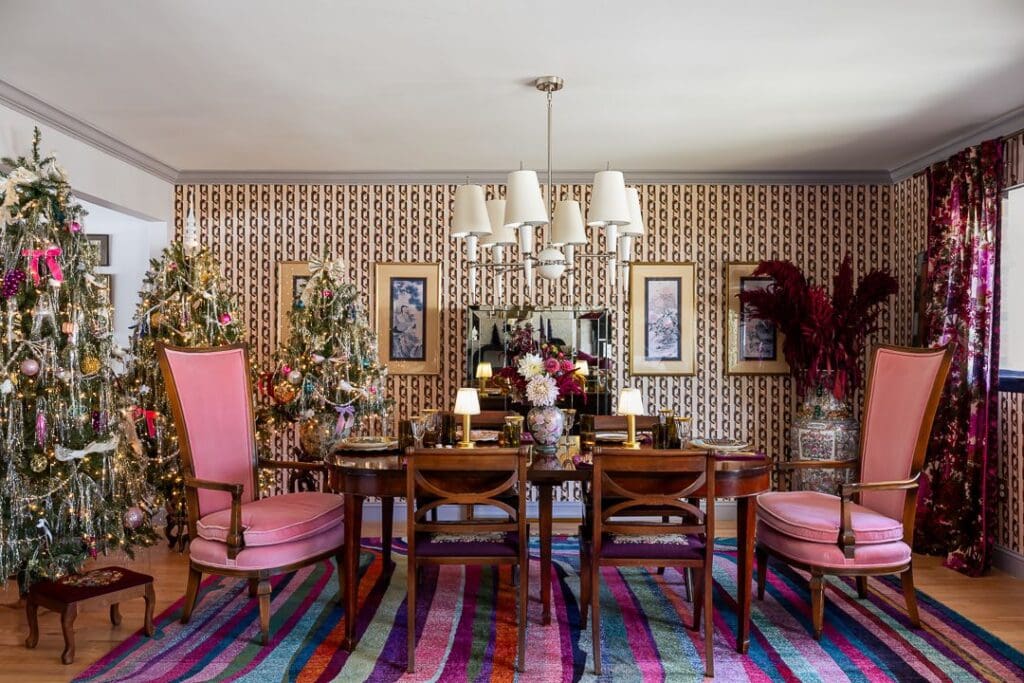 A vibrant holiday dining room styled with bold vintage charm. Pink high-back armchairs flank a wooden dining table set with floral china, gold chargers, and mid-century lamps
