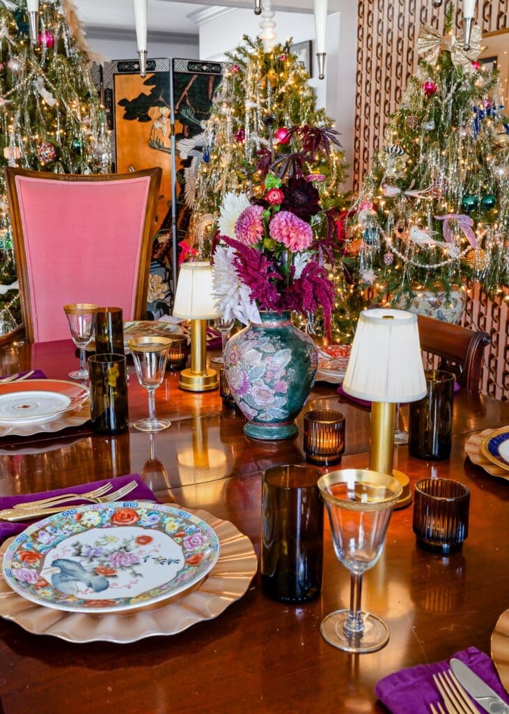 A richly set holiday dining table featuring layered vintage china, gold-rimmed glassware, amber votives, and petite brass table lamps. A lush floral arrangement in a decorative vase anchors the centerpiece, while three tinsel-adorned Christmas trees sparkle in the background, creating a maximalist yet refined holiday ambiance.