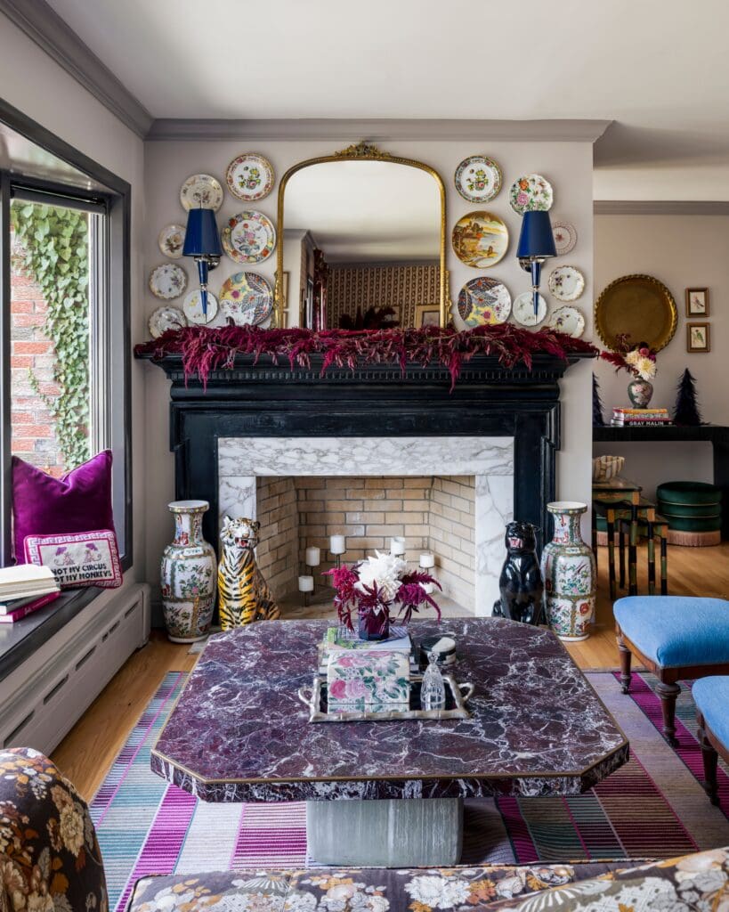 Eclectic holiday living room styled with vintage china, a marble fireplace draped in burgundy garland, and layered jewel-toned textiles. A gold mirror and sculptural decor frame the space, blending traditional charm with maximalist flair