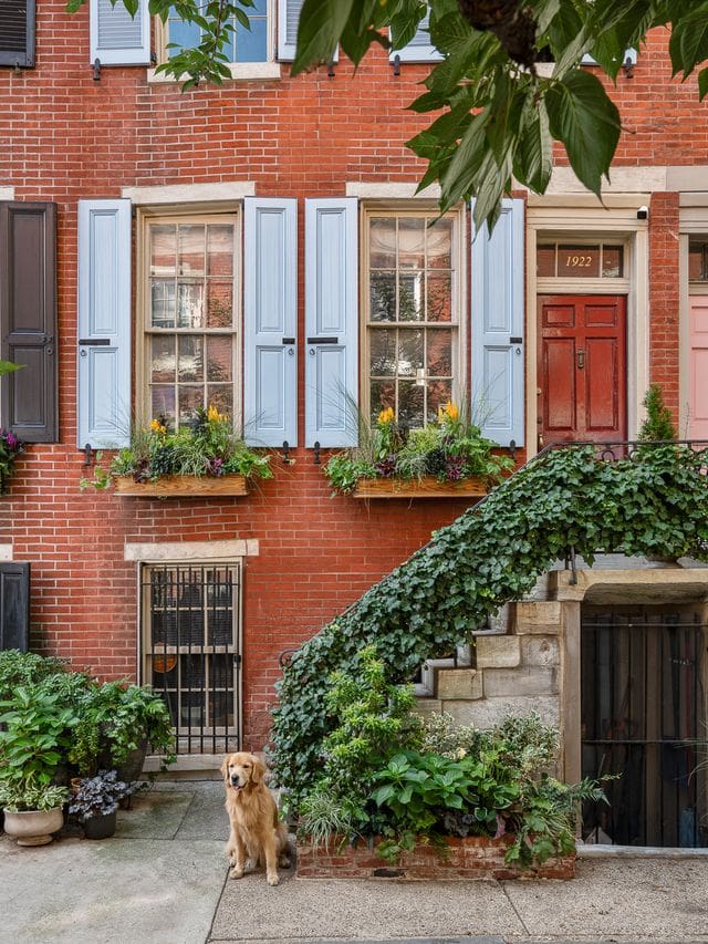 A golden retriever sits in front of a red-brick townhouse adorned with lush green plantings and soft blue shutters, captured in natural light. A warm editorial image highlighting community, design, and charm.