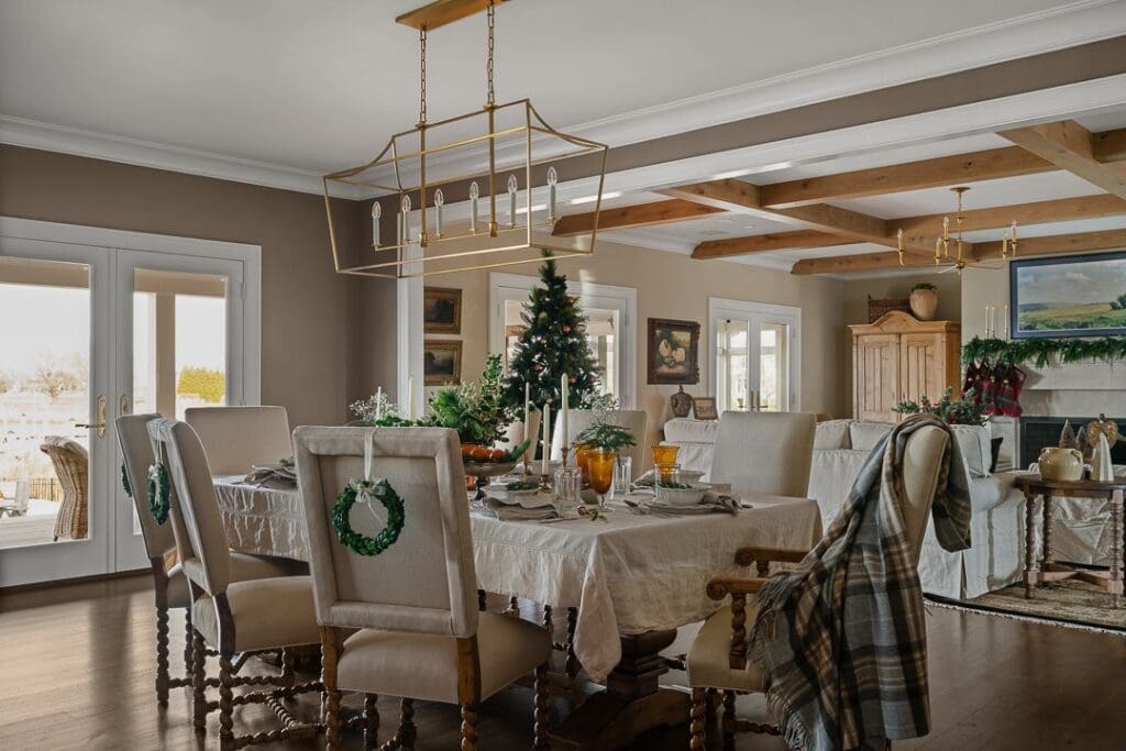 A welcoming dining room with a linen-covered table, small evergreen wreaths on chair backs, and warm brass lighting overhead. The scene feels festive yet effortlessly relaxed.