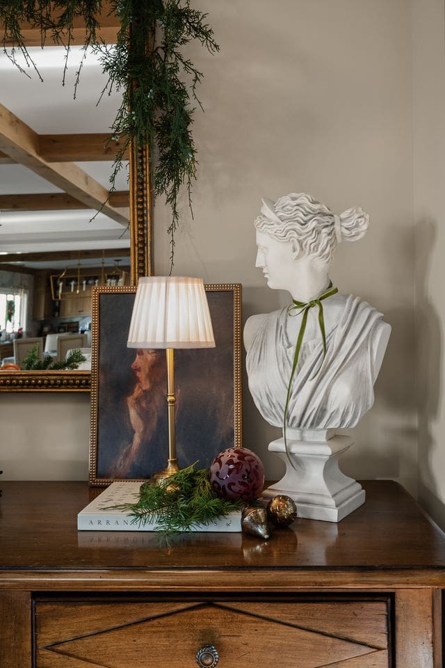 A sculptural holiday vignette featuring a classical bust adorned with a velvet ribbon, set beside a moody portrait, fluted brass lamp, and vintage ornaments. Captured by architectural digest approved photographer Tori Sikkema.
