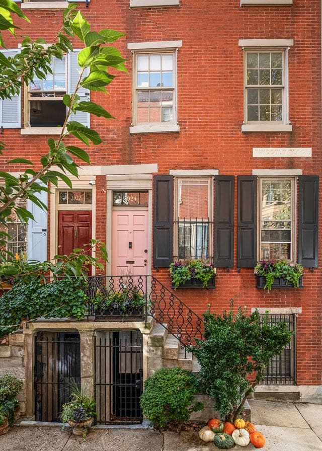 Pink and red townhouse doors framed by layered greenery, black shutters, and handcrafted floral window boxes, styled by Hannah Levine. A refined expression of quiet luxury and neighborhood connection.