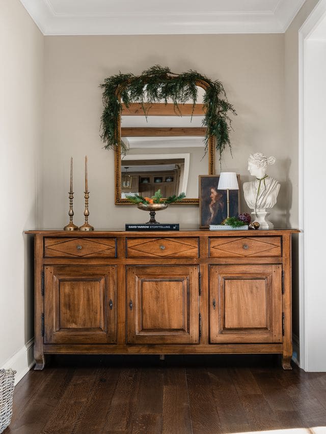 A refined vignette of a heritage-inspired console table styled for the holidays, featuring quiet luxury details. The warm wood tones of the antique buffet are complemented by layered holiday greenery draped over a gilt-framed mirror.