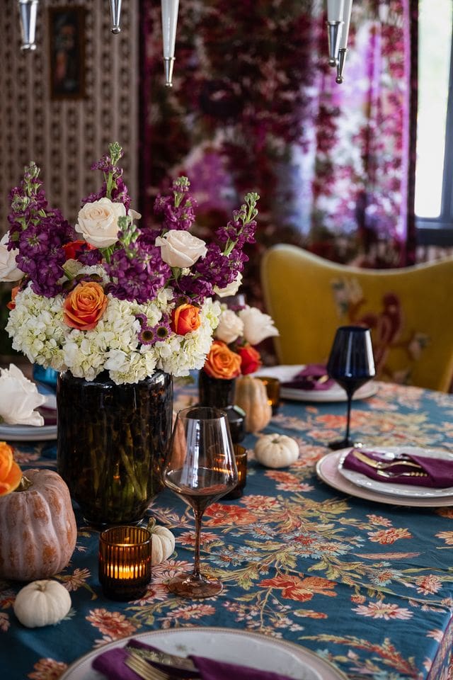 Lush floral centerpieces anchor the composition, surrounded by white mini pumpkins, evoking a cozy yet curated fall gathering. The vibrant floral curtain and embroidered yellow dining chairs add depth and dimension to the scene, blending vintage maximalism with editorial sophistication.