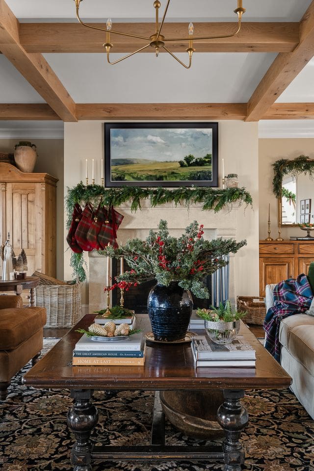 A warm living room with a garland-draped mantel, plaid stockings, and a large floral arrangement of evergreens and berries in a dark ceramic vase. Stacked design books and soft plaid throws evoke timeless holiday comfort.