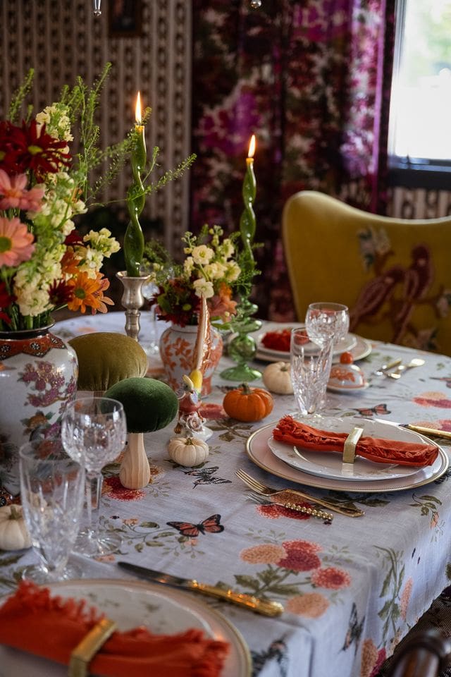 The scene evokes editorial elegance, maximalist decor, and festive tabletop styling perfect for a holiday or autumn gathering photographed by AD Approved Tori Sikkema. A close-up of an opulent fall tablescape featuring layered floral china, crystal stemware, and playful green taper candles in vintage holders.