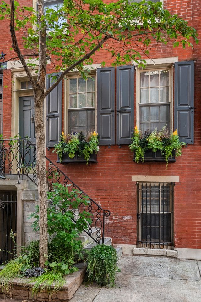 A red-brick Philadelphia townhouse with charcoal shutters and lush window boxes designed by Hannah Levine of Flornamentum, featuring cascading greenery and golden blooms. Captured by AD-Approved interior photographer Tori Sikkema, showcasing floral design that enhances historic architecture.