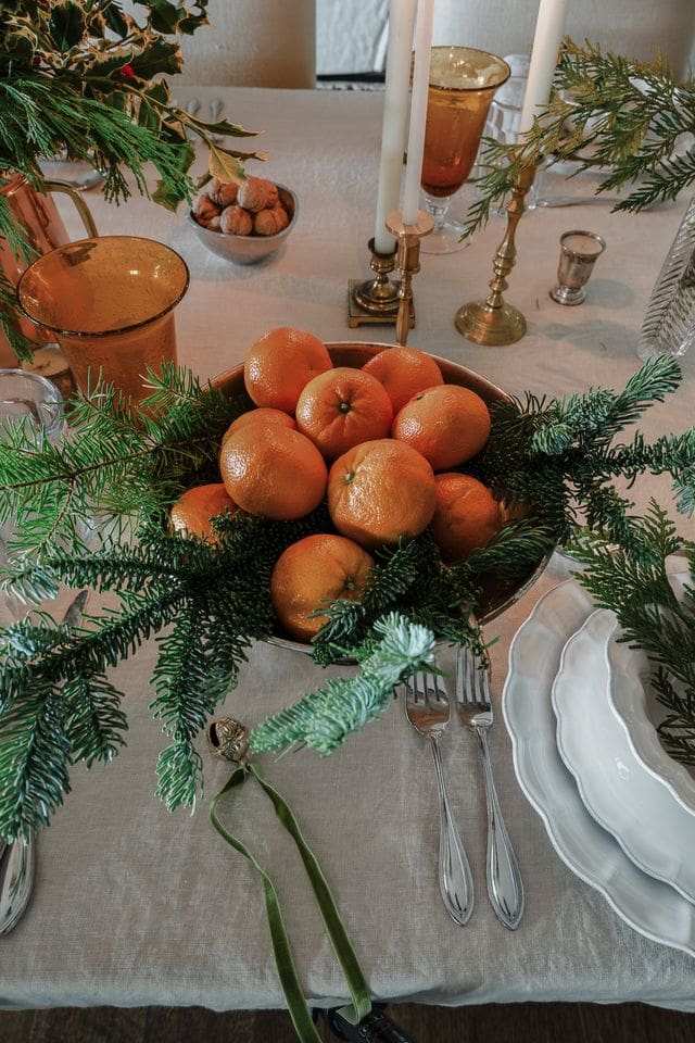 A simple bowl of tangerines nestled in fir branches and tied with a green velvet ribbon adds a natural, joyful touch to the holiday table.