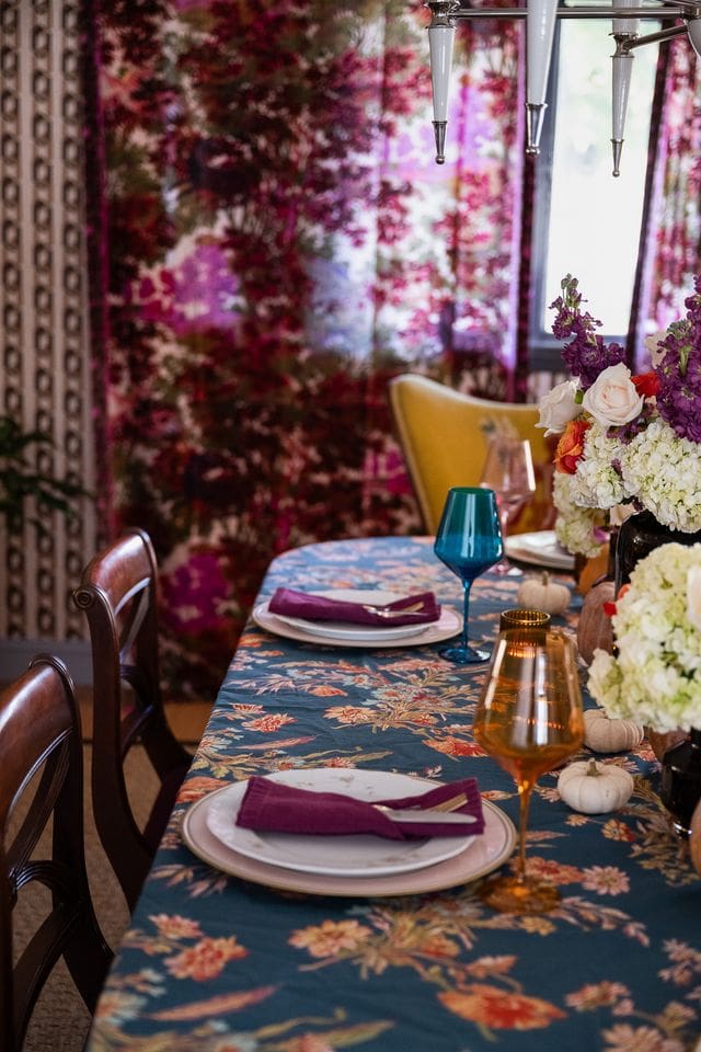 A moody editorial photograph of a floral tablescape with jewel-toned accents. Lush floral centerpieces anchor the composition, surrounded by white mini pumpkins, evoking a cozy yet curated fall gathering. 