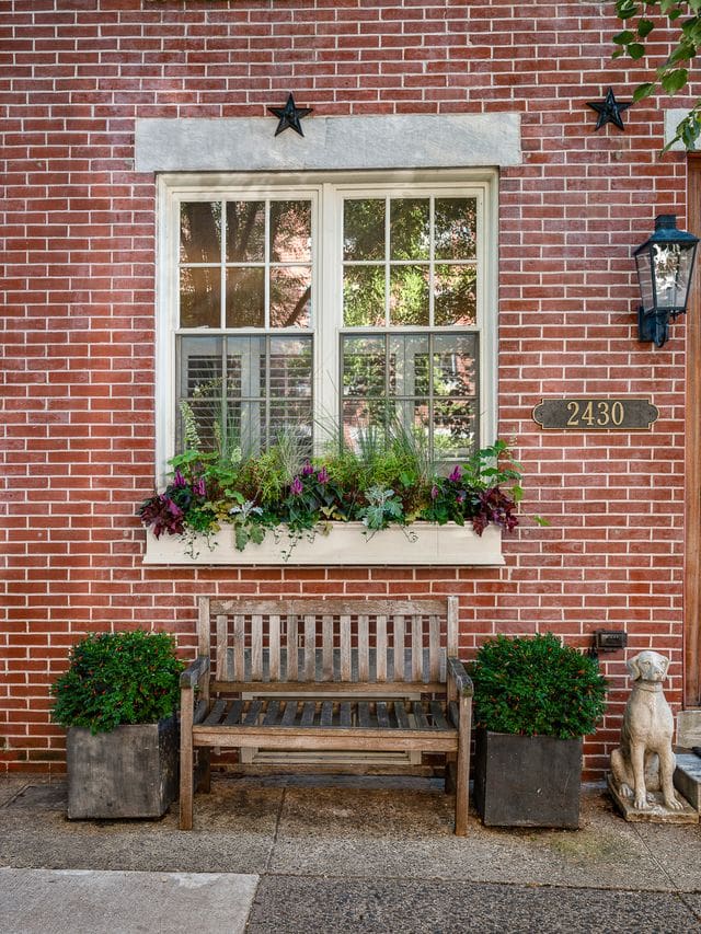 Brick façade with a natural wood front door, black shutters, and a single flower box overflowing with purple and green foliage. Pumpkins and a stone dog statue add charm to this classic Philadelphia home.