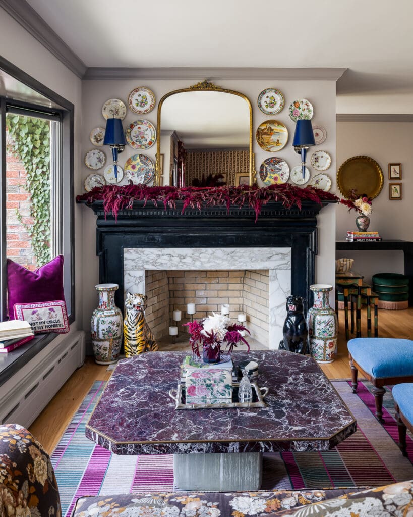 Retro-inspired living room with tapestry-style floral sofa, bright blue slipper chairs, marble coffee table, striped rug, and adjoining view into a bold patterned dining room.