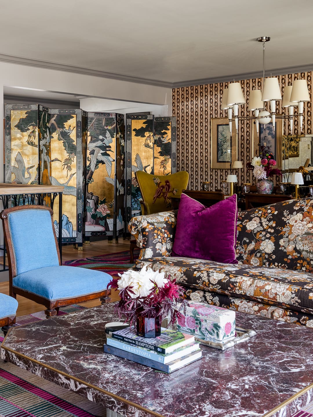 Retro-inspired living room with tapestry-style floral sofa, bright blue slipper chairs, marble coffee table, striped rug, and adjoining view into a bold patterned dining room.