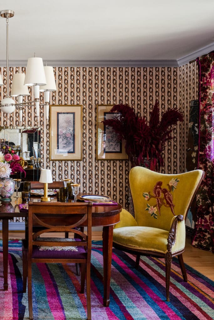 Pattern-rich retro dining room with graphic wallpaper, embroidered chartreuse chairs, dark wood dining table, colorful striped rug, and layered vintage art and décor.