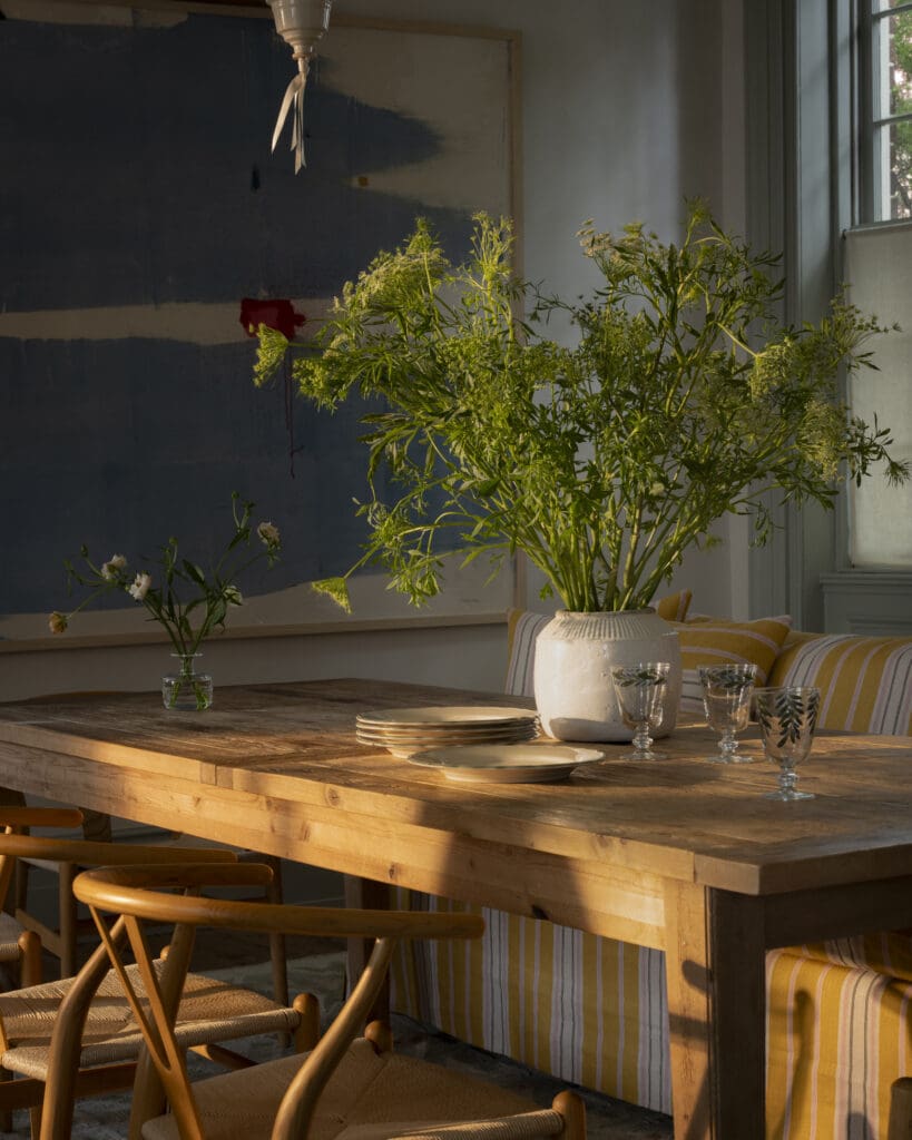 Warmly lit dining room designed by Kate Towill in Charleston, photographed by Tori Sikkema. The space features a rustic wood dining table, classic Wishbone chairs, a striped slipcovered sofa, and a bold abstract painting. A whimsical oversized pendant light with red trim anchors the room, while natural light pours in through paned windows, casting soft shadows across the vintage rug and wide plank wood floors.