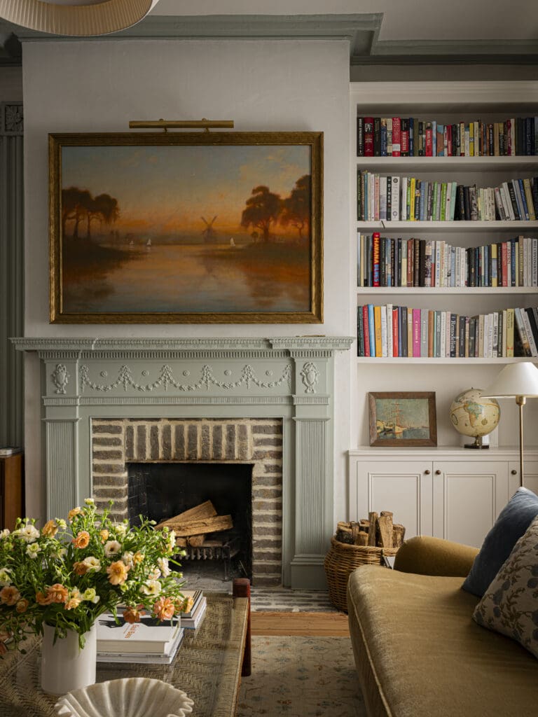 A layered living room with a historic brick fireplace, mint-green mantel, and a sunset landscape painting, styled with vintage accents, a curated bookshelf, and fresh florals. Interior design by Kate Towill. Photo by Tori Sikkema Photography