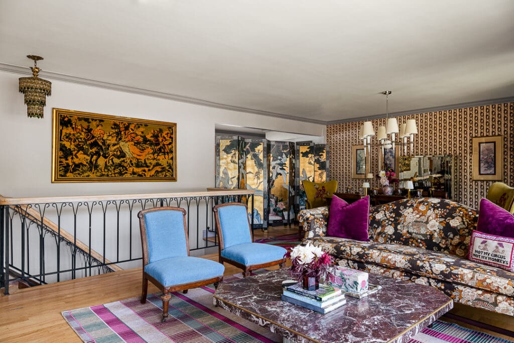 Retro-inspired living room with tapestry-style floral sofa, bright blue slipper chairs, marble coffee table, striped rug, and adjoining view into a bold patterned dining room.