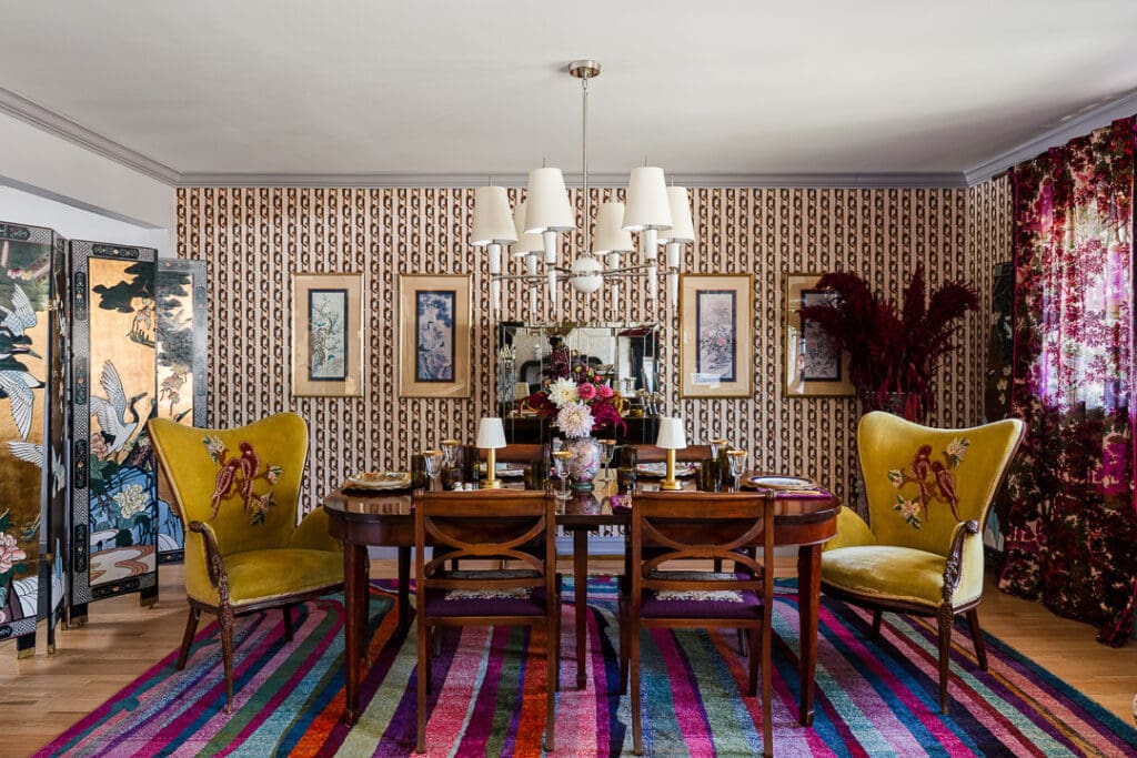 Pattern-rich retro dining room with graphic wallpaper, embroidered chartreuse chairs, dark wood dining table, colorful striped rug, and layered vintage art and décor.